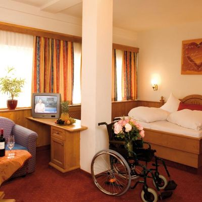 The image shows a wheelchair in the middle of a room with flowers on it. To the left is a table with two glasses of wine next to a couch and a TV. To the right, behind the chair, is a bed, above which hangs a picture of a heart.