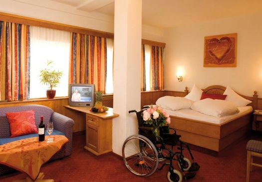 The image shows a wheelchair in the middle of a room with flowers on it. To the left is a table with two glasses of wine next to a couch and a TV. To the right, behind the chair, is a bed, above which hangs a picture of a heart.